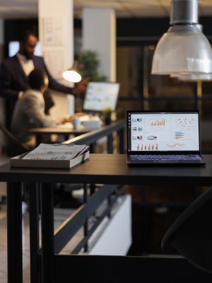 Laptop computer with financial graphs on screen Laptop computer with financial graphs on screen, standing on table in startup office, coworkers discussing company strategy in background. Employees working late at night in startup office